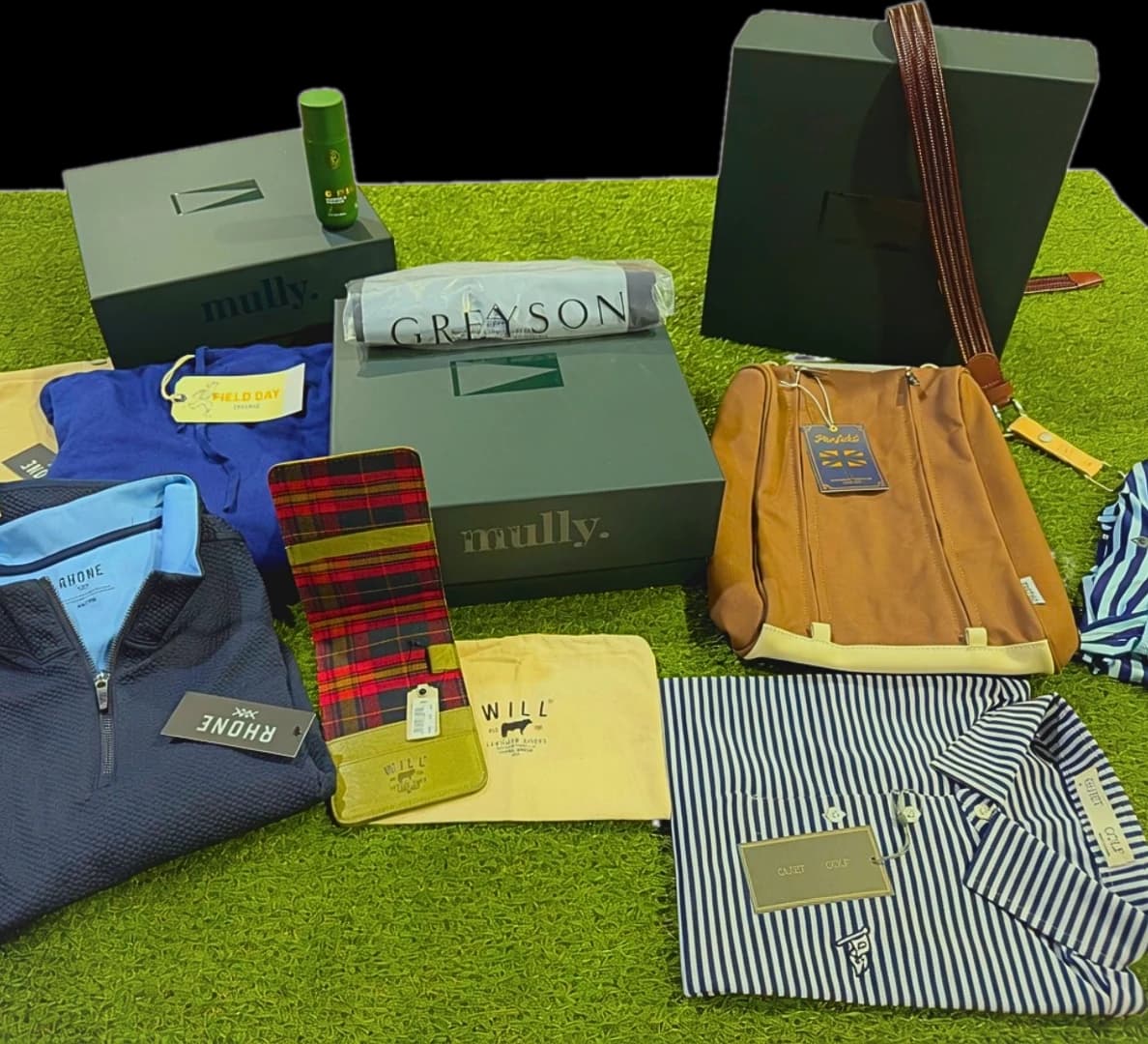 An editor's flat-lay of a recent Mully Reserve box on turf — Greyson tee, Rhone quarter-zip, Field Day hoodie, Will Leather goods, Penfold shoe bag, Cuater polo, and a Mully box.